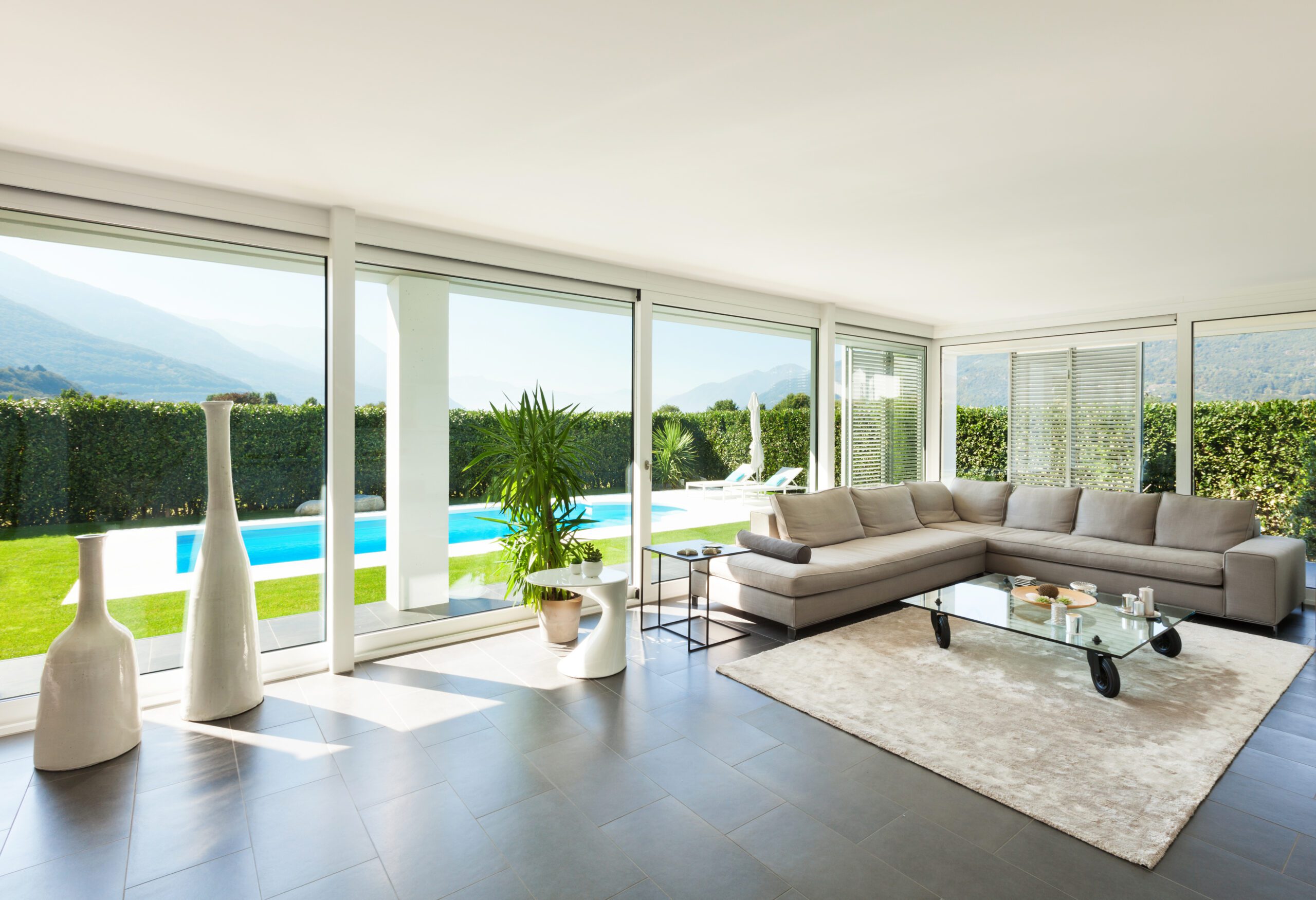 Modern living room with large windows overlooking a backyard with a pool and mountain view. Features a gray sectional sofa, glass coffee table, potted plants, and white vases.