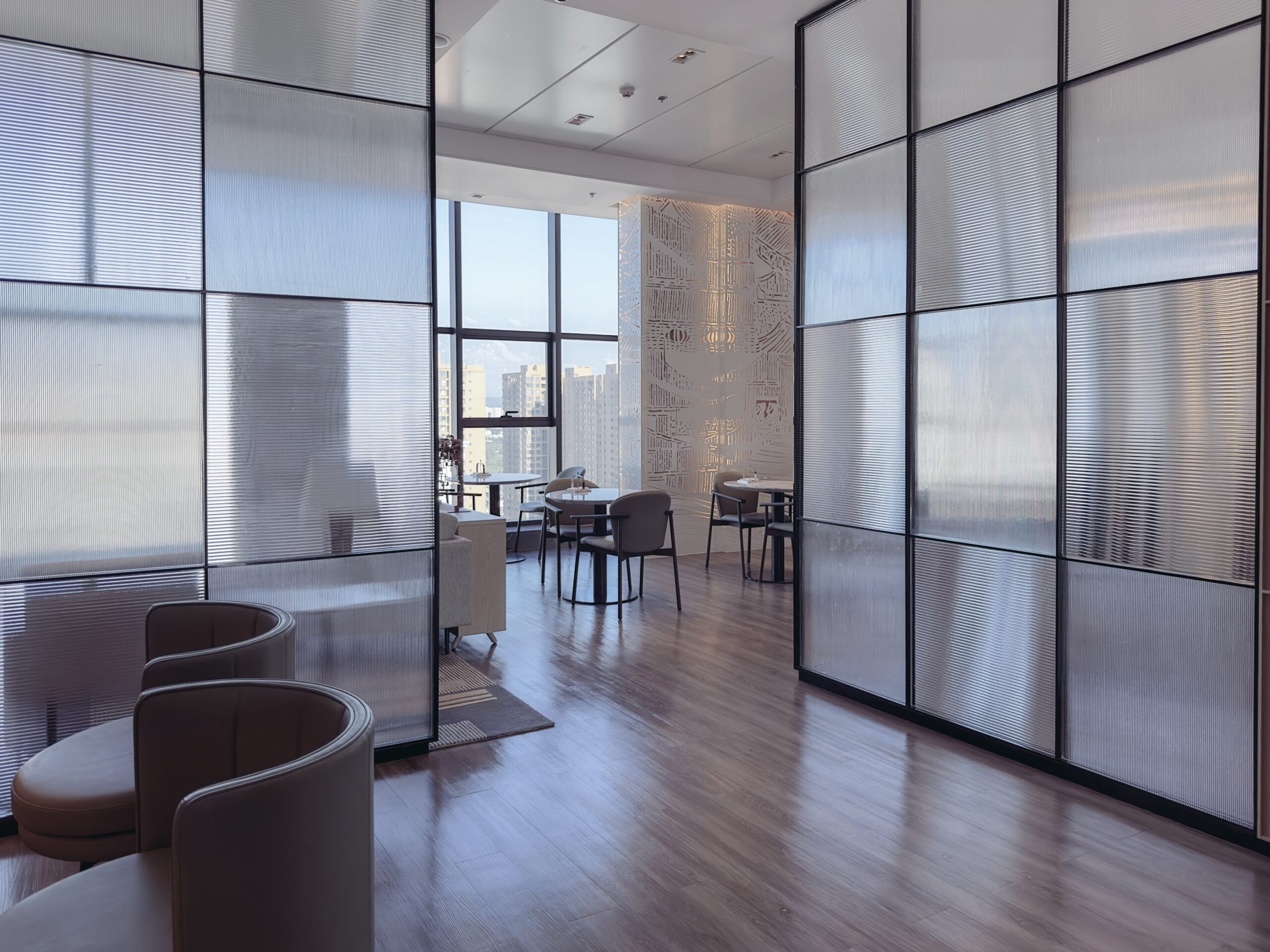 Modern office lounge with floor-to-ceiling windows, minimalistic furniture, wooden flooring, and divided sections by transparent partitions. Wall art is visible in the background.